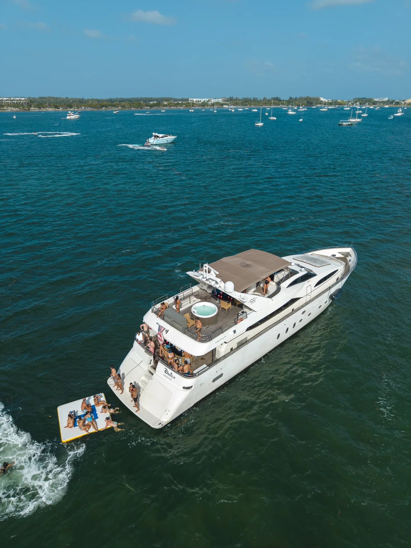 Aerial drone photo of a luxury motor yacht anchored on Biscayne Bay near Miami, with guests on a floating water mat and swimmers in turquoise water