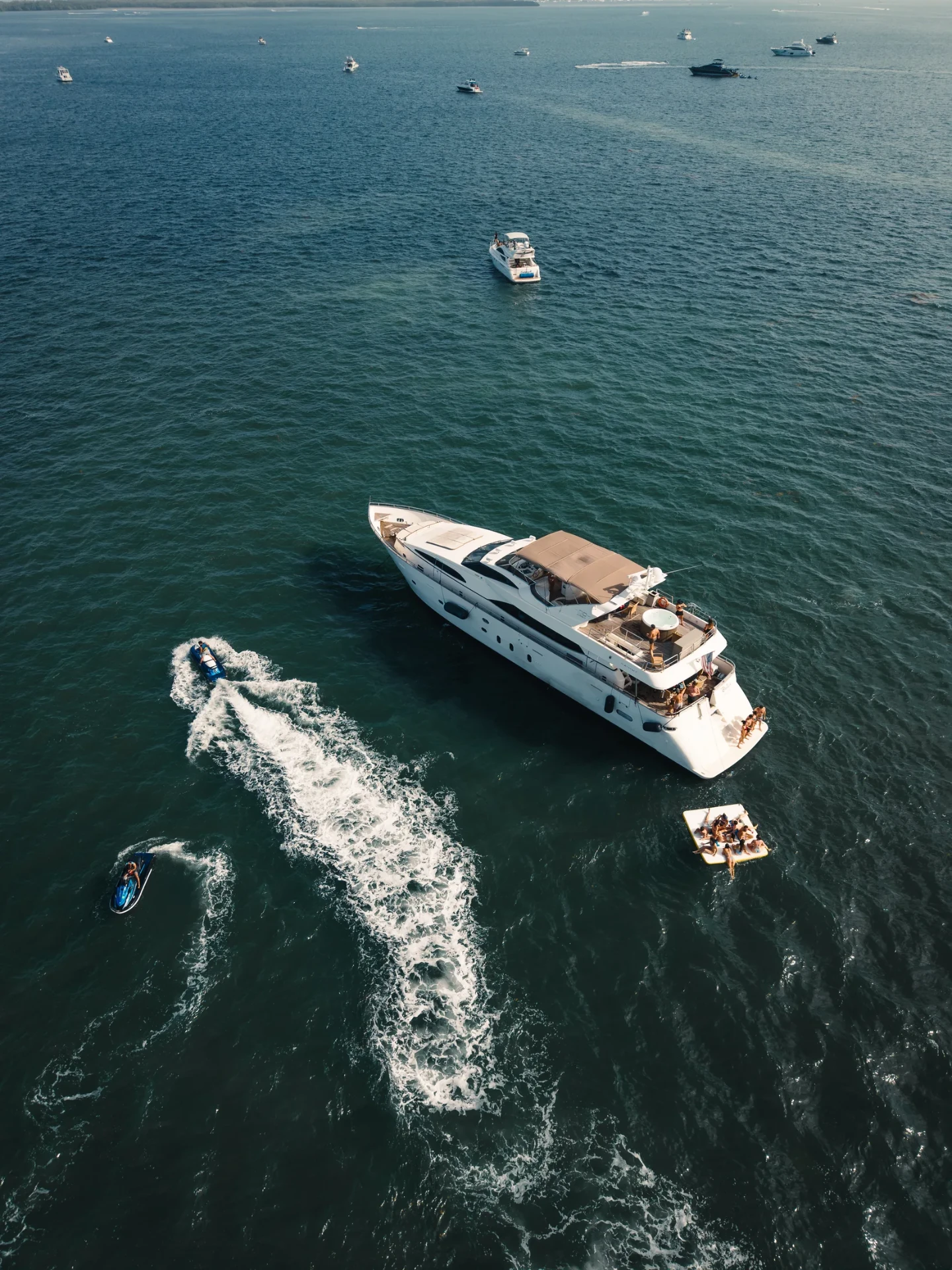 Aerial drone view of a luxury yacht with guests aboard and jet skis carving through the water off Miami